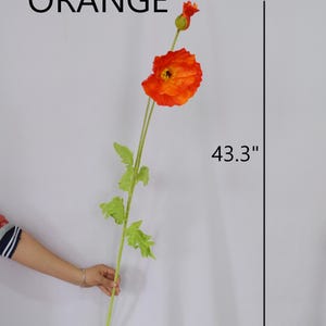 May include: An orange artificial poppy flower with green leaves and stem. The flower is 43.3 inches long and has a vibrant orange color. The word "ORANGE" is written in black letters above the flower.