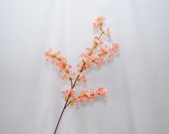 May include: A decorative artificial branch featuring numerous small, delicate peach-colored blossoms and light green leaves attached to a thin, dark brown stem. The branch extends diagonally across the frame, showcasing multiple clusters of flowers.