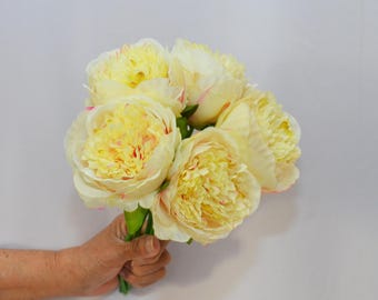May include: A bouquet of artificial peonies in shades of yellow and cream, with hints of pink on the edges of the petals. The flowers are held together by green stems, creating a vibrant floral arrangement.