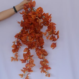 May include: A long strand of artificial fall foliage with orange and brown leaves. The leaves are made of a thin, plastic material and are arranged in a cascading pattern.