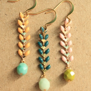 May include: Three pairs of gold dangle earrings with a chevron pattern. The earrings are in three different color combinations: yellow and light blue, teal and gold, and pink and gold. Each earring has a small round bead at the bottom.