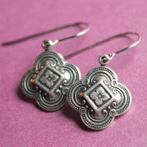 May include: A pair of silver dangle earrings with a floral design. The earrings have a four-leaf clover shape with a small flower in the center of each.