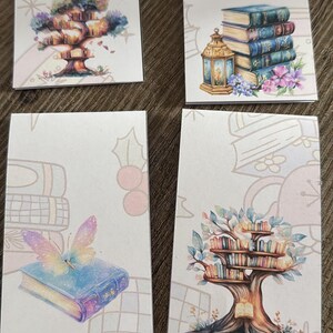 Könnte beinhalten: Vier rechteckige Karten mit skurrilen Illustrationen. Eine zeigt ein baumförmiges Bücherregal, eine andere einen Stapel Bücher mit einer Laterne, eine dritte ein Buch mit einem Schmetterling und die letzte ein baumförmiges Bücherregal. Jede Karte hat einen hellen Hintergrund mit floralen und Schneeflocken-Akzenten.