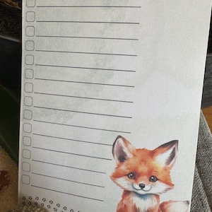 Könnte beinhalten: Ein Notizblock mit dem Text "Meine FUCHStastische Einkaufsliste" oben. Der Notizblock hat eine niedliche Cartoon-Fuchs-Illustration unten rechts. Der Fuchs ist orange, weiß und braun. Der Notizblock hat Linien zum Schreiben.