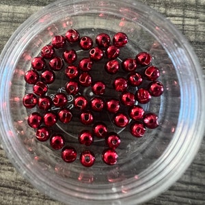 Könnte beinhalten: Nahaufnahme eines durchsichtigen Plastikbehälters gefüllt mit roten Glasperlen. Die Perlen sind rund und haben eine glänzende Oberfläche. Jede Perle hat einen eingeprägten Buchstaben.
