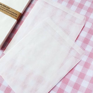 May include: A stack of white paper envelopes on a pink and white gingham tablecloth. The envelopes are made from 100% pine cellulose and are compostable. The text on the envelope reads "etsy.cicia.com UK from 100% pine cellulose. Dispose of it in a compost bin."