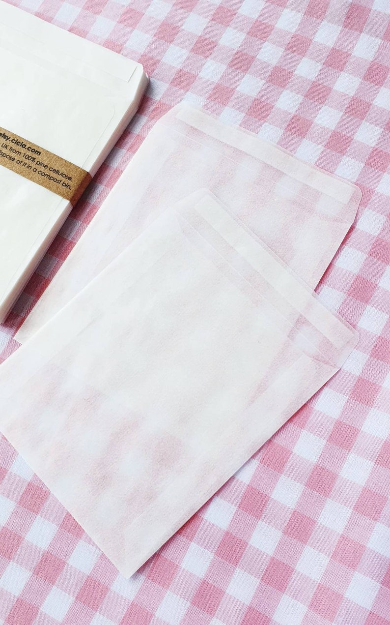 May include: A stack of white paper envelopes on a pink and white gingham tablecloth. The envelopes are made from 100% pine cellulose and are compostable. The text on the envelope reads "etsy.cicia.com UK from 100% pine cellulose. Dispose of it in a compost bin."