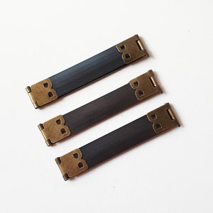 Könnte beinhalten: Drei rechteckige, dunkelbraune Metallscharniere mit antiken messingfarbenen Eckwinkeln. Die Scharniere sind diagonal auf einer weißen Oberfläche angeordnet, ideal für Holzbearbeitungs- oder Bastelprojekte.