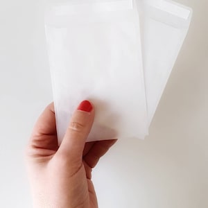 May include: Two white paper envelopes with a sealed flap. The envelopes are held in a hand with red nail polish.