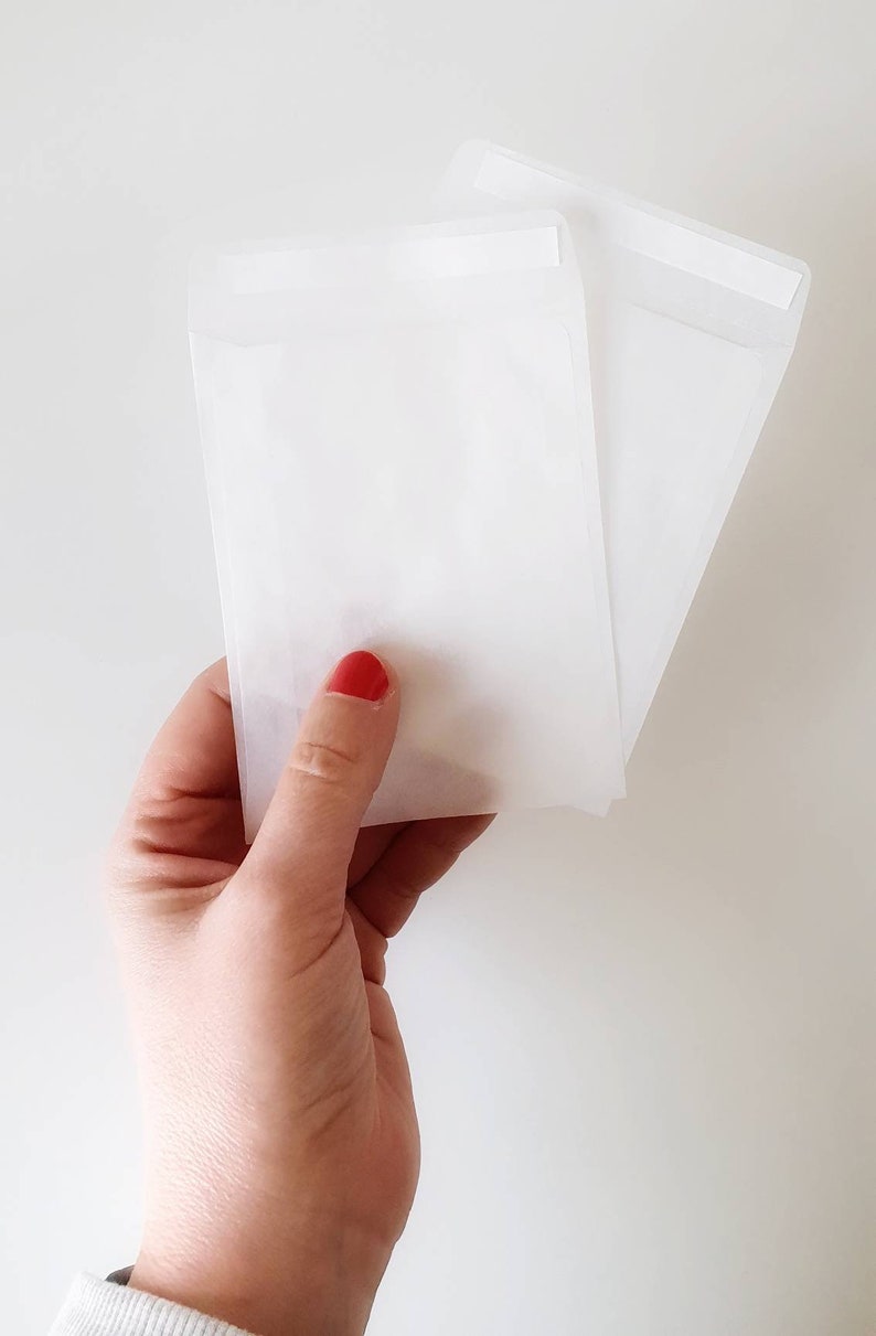 May include: Two white paper envelopes with a sealed flap. The envelopes are held in a hand with red nail polish.