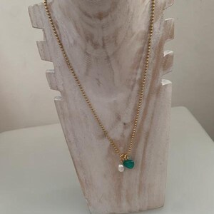 May include: A gold-colored necklace with a delicate chain, featuring a small green heart-shaped charm and a white pearl pendant. The necklace is displayed on a light-colored wooden jewelry stand.