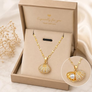 May include: A gold-toned necklace featuring a shell-shaped pendant encrusted with clear stones, presented in a beige box with the text "Especially for you Made with love". An inset shows the pendant opened to reveal a white spherical pearl inside.