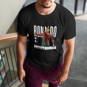 May include: A black t-shirt with a white graphic of Cristiano Ronaldo celebrating a goal. The graphic includes the text "RONALDO" and "7."  The graphic also includes a row of five soccer trophies.