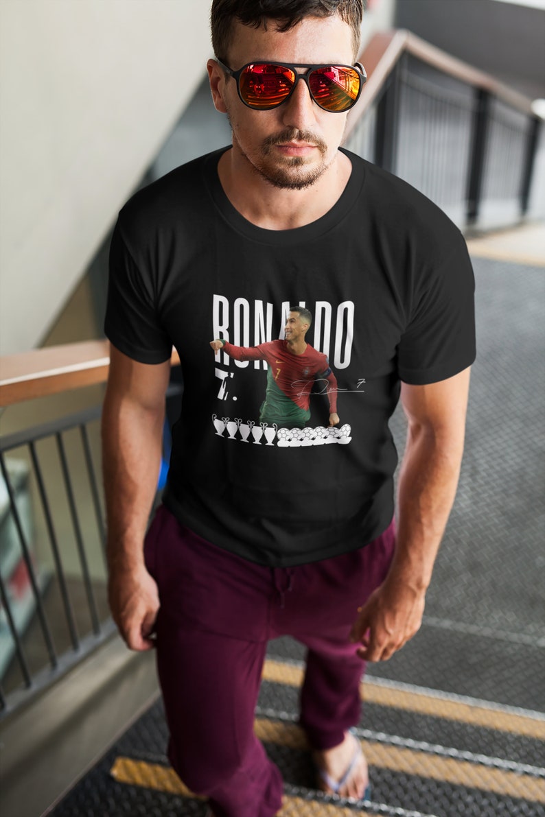 May include: A black t-shirt with a white graphic of Cristiano Ronaldo celebrating a goal. The graphic includes the text "RONALDO" and "7."  The graphic also includes a row of five soccer trophies.