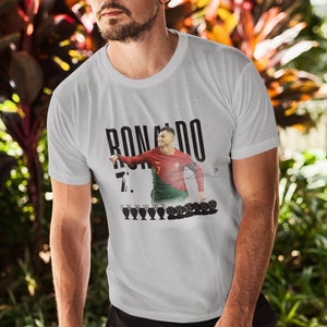 May include: White t-shirt with a black and white image of Cristiano Ronaldo in a Portugal jersey. The text "RONALDO 7." is printed on the shirt. There are five black and white soccer trophies and several soccer balls printed below the image.