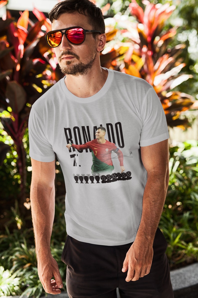May include: White t-shirt with a black and white image of Cristiano Ronaldo in a Portugal jersey. The text "RONALDO 7." is printed on the shirt. There are five black and white soccer trophies and several soccer balls printed below the image.