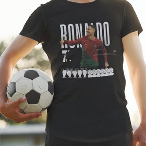 May include: Black t-shirt with a white graphic of Cristiano Ronaldo in a soccer uniform, the text "RONALDO 7." and a signature. The graphic also includes a row of soccer balls and trophies.
