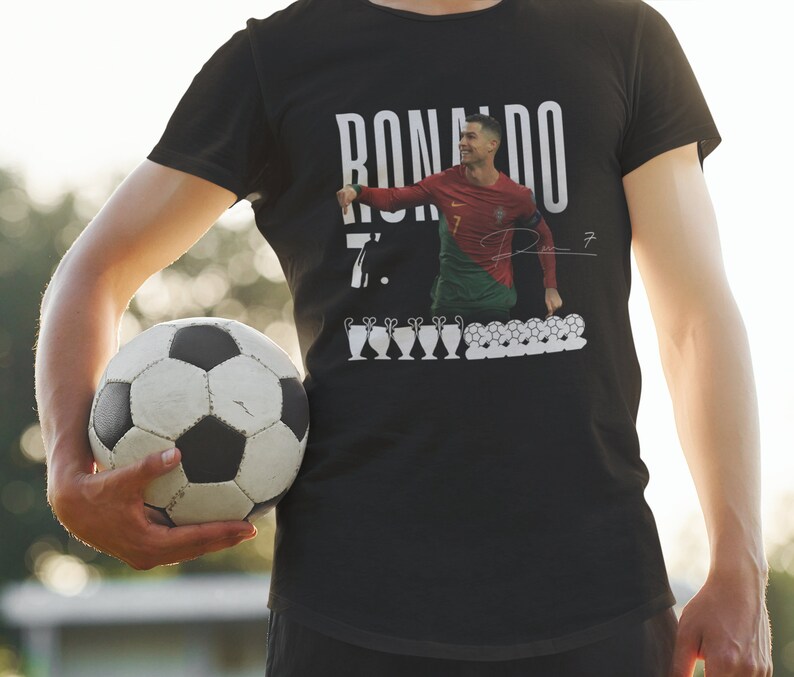 May include: Black t-shirt with a white graphic of Cristiano Ronaldo in a soccer uniform, the text "RONALDO 7." and a signature. The graphic also includes a row of soccer balls and trophies.
