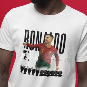 May include: White t-shirt with a black and white image of Cristiano Ronaldo in a soccer uniform. The text "RONALDO 7." is printed on the shirt. There are also images of soccer balls and trophies.