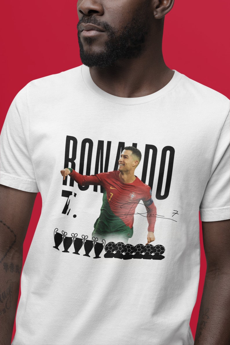 May include: White t-shirt with a black and white image of Cristiano Ronaldo in a soccer uniform. The text "RONALDO 7." is printed on the shirt. There are also images of soccer balls and trophies.