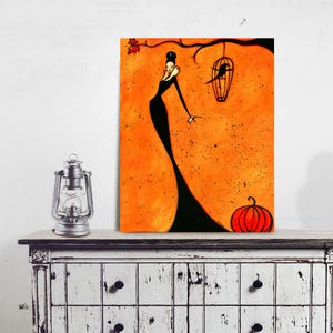 May include: An orange and black art print featuring a woman in a long black dress, a bird in a cage, and a pumpkin. The artwork is displayed on a distressed white cabinet with a silver lantern and a dress form.