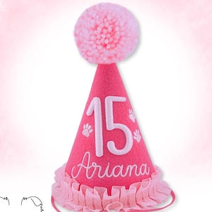 May include: Pink felt party hat with a white pom-pom on top. The hat has a white number "15" and the name "Ariana" embroidered on it. There are also white paw prints embroidered on the hat.