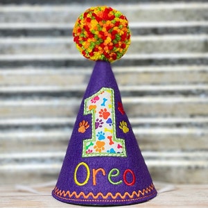 May include: Purple felt party hat with a colorful pom-pom on top. The hat has a white felt patch with a number one, paw prints, and bone designs. The name "Oreo" is embroidered on the hat.