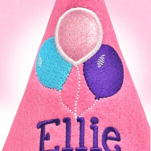 Op de afbeelding: Roze feesthoed met geborduurde ballonnen in blauw, roze en paars. De naam "Ellie" is eronder in paars geborduurd. De hoed is van vilt.