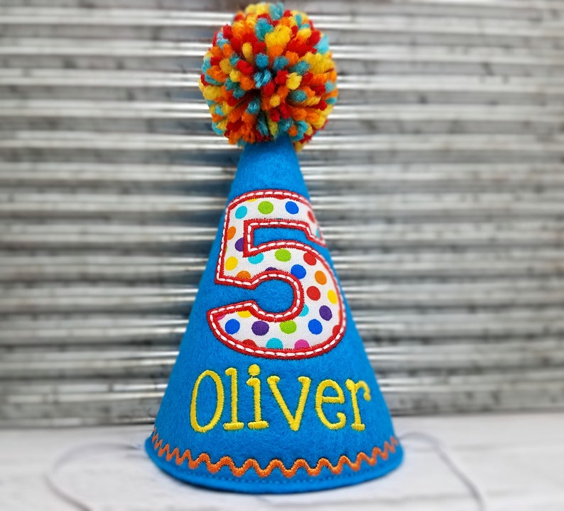 May include: A blue birthday party hat with a colorful pom-pom on top. The number "5" is displayed on the front with a polka dot pattern. The name "Oliver" is embroidered in yellow. The hat has an orange zig-zag trim.
