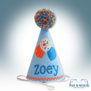 May include: Blue felt party hat with a white pom pom on top. The hat has a colorful balloon design and the name "Zoey" embroidered on the front.
