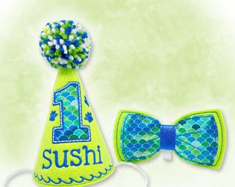 May include: A lime green party hat with a blue, green, and white pom-pom, featuring a blue number 1 and the name Sushi in blue embroidery, alongside a matching blue and green scale-patterned bow tie with a blue center strap.