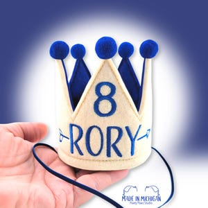May include: A beige felt crown featuring a royal blue embroidered number 8 and the name RORY flanked by two blue arrows. The crown is topped with five blue pom-poms and includes a thin blue elastic chin strap for secure placement.