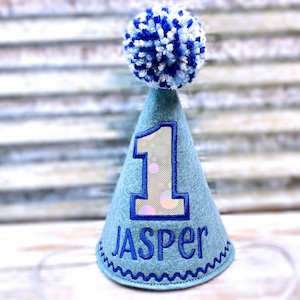 May include: Blue felt party hat with a white pom pom on top. The hat has a large number "1" with a glittery background and the name "Jasper" embroidered on the front.