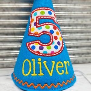 May include: A blue felt birthday party hat with a large number 5 in the center, outlined in red and filled with colorful polka dots. The name "Oliver" is embroidered in yellow below the number. The hat has an orange zig-zag trim.