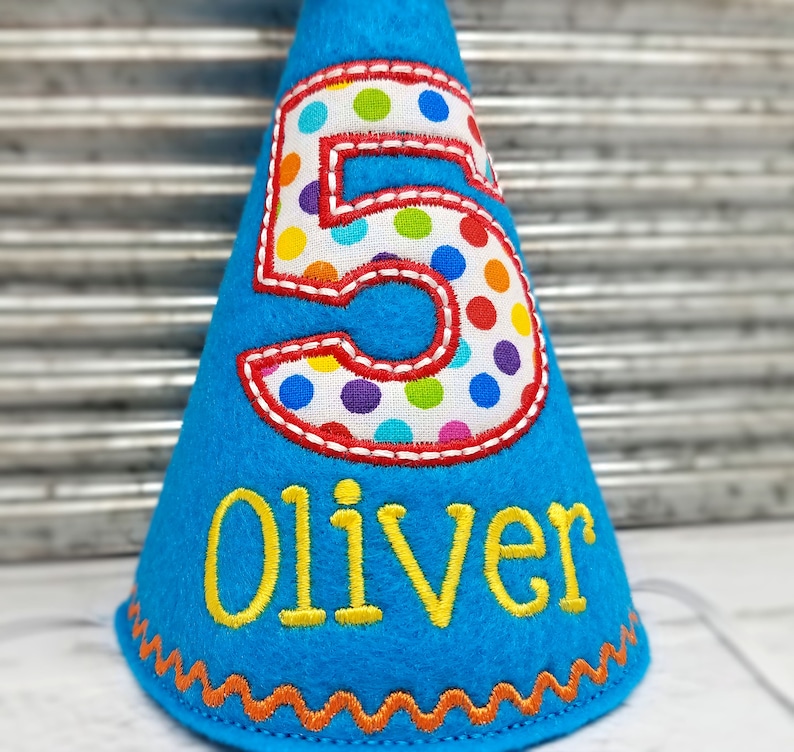 May include: A blue felt birthday party hat with a large number 5 in the center, outlined in red and filled with colorful polka dots. The name "Oliver" is embroidered in yellow below the number. The hat has an orange zig-zag trim.