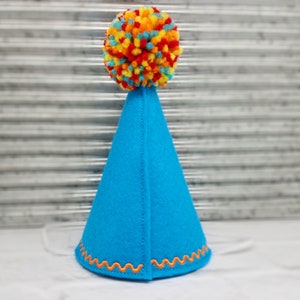 May include: A bright blue party hat with an orange zig-zag trim and a colorful pom-pom on top. The hat is made of felt and has a white string to secure it. This festive hat is perfect for celebrations.