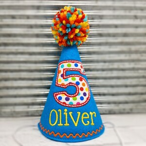 May include: A blue birthday party hat with a colorful pom-pom on top. The number "5" is appliqued on the front with a polka dot pattern. The name "Oliver" is embroidered in yellow. The hat has an orange zig-zag trim.