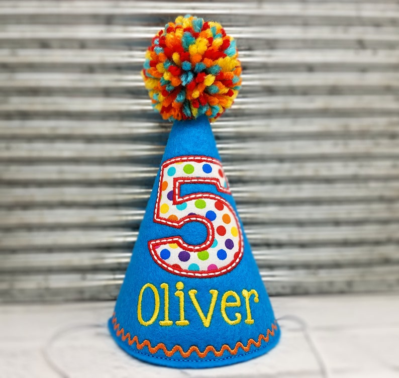 May include: A blue birthday party hat with a colorful pom-pom on top. The number "5" is appliqued on the front with a polka dot pattern. The name "Oliver" is embroidered in yellow. The hat has an orange zig-zag trim.
