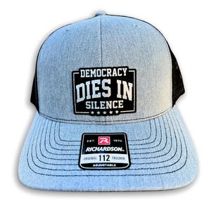 May include: A grey and black trucker hat with a black patch that reads "DEMOCRACY DIES IN SILENCE" with five stars below the text. The hat has a black mesh back and a red and white label that reads "RICHARDSON. ORIGINAL 112 TRUCKER ADJUSTABLE".