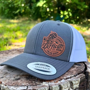 Op de afbeelding: Een grijze en witte truckercap met een bruin leren patch waarop staat "Duur en moeilijk, praat terug". De patch toont een schedel met een bliksem en een bierpul. De pet heeft een YUPOONG-logo en er staat "The Authentic Snapback, YUPOONG Since 1974" op.