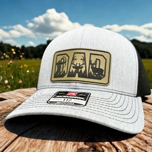 Pode incluir: Boné de caminhoneiro cinza claro com parte traseira em tela preta. A frente apresenta um patch com a palavra "DAD" e silhuetas de um caçador, veado e árvores. O boné tem uma aba curva e uma alça ajustável.
