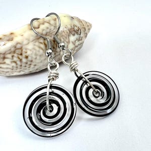 May include: A pair of spiral design earrings. Each earring features a black and white spiral pattern within a clear, circular frame. The earrings are suspended from silver-colored hooks.