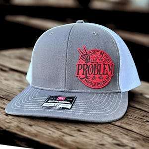 May include: Grey and white trucker cap featuring a red patch with the text "DON'T BE PART Of The PROBLEM Be The WHOLE PROBLEM". The cap has a curved peak and an adjustable strap.