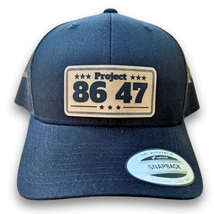 May include: Black trucker hat with a brown leather patch that says "Project 86 47" with stars around the text. The hat has a black mesh back and a black snap closure.