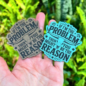 Puede incluir: Dos pegatinas con temática del oeste. Una es de color canela, la otra turquesa. Ambas tienen el texto "Si yo soy el problema, entonces tú podrías ser la razón" con un gráfico de calavera de toro. Las pegatinas se sostienen en una mano.
