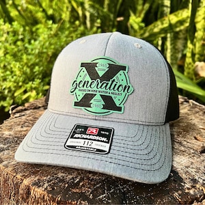 Puede incluir: Gorra de camionero gris y negra con visera curva. La gorra presenta un parche con la palabra "generation" y los años 1965 y 1980. La gorra es ajustable y tiene la etiqueta de la marca Richardson.