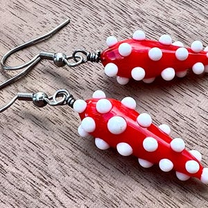 May include: A pair of red and white glass earrings with a teardrop shape and white polka dots. The earrings are attached to silver hooks.