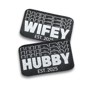 Puede incluir: Dos parches de cuero negro con texto blanco. El parche superior dice "WIFEY EST. 2025" y el parche inferior dice "HUBBY EST. 2025".
