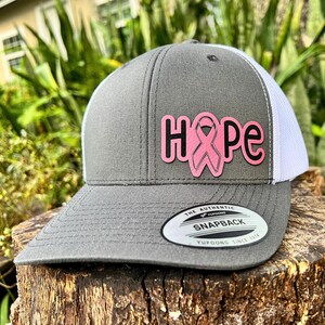 May include: A gray and white trucker hat with the word "HOPE" in pink letters and a pink ribbon graphic. The hat has a snapback closure and a curved brim. The hat is made by Yupoong.