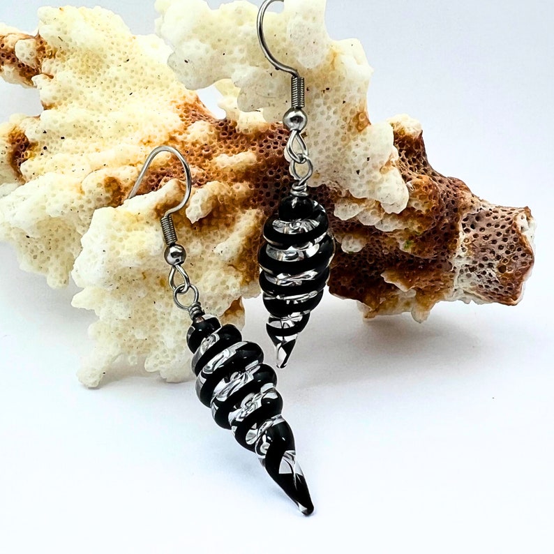 May include: A pair of black and silver spiral earrings with silver-colored ear wires. The earrings are cone-shaped and have alternating black and silver bands. The earrings are displayed in front of a piece of white coral.